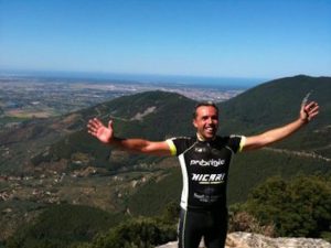 Billede af cykelrytter i Italien. Cykkelrytteren står med ryggen til smuk italiensk natur og bjerge. Cykling i Italien.